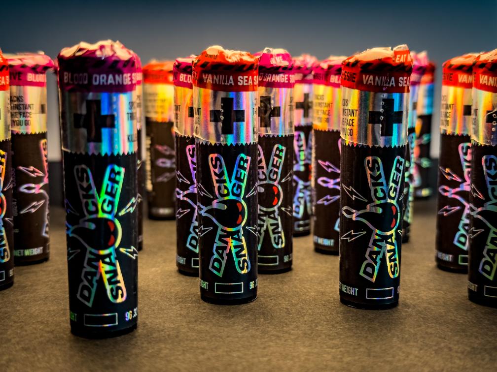 Danger Snacks Fire Pellets tubes with holographic labels, featuring Blood Orange Sours and Vanilla Sea Salt flavors arranged on a surface.